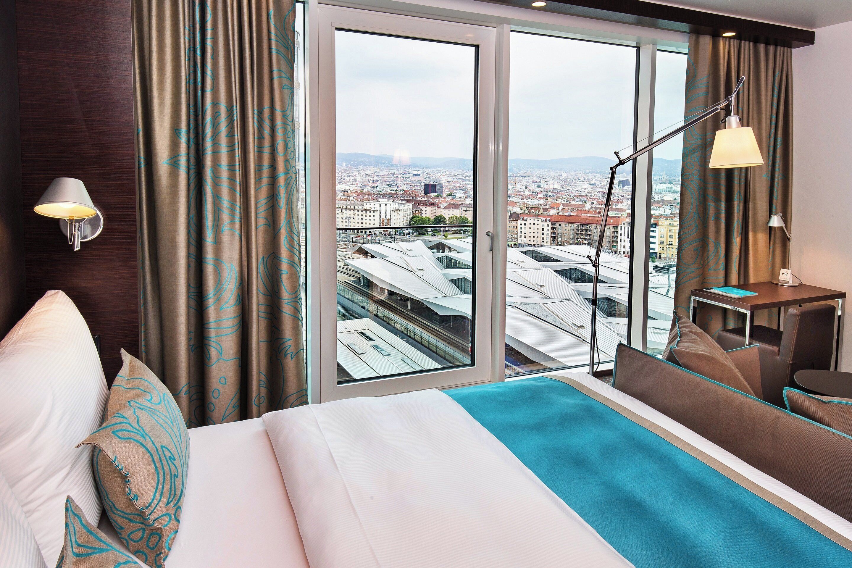 A hotel room with a large bed, a desk, a chair, a standing lamp, and a view of the city through a large window.