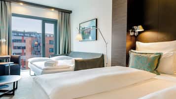 A hotel room with a large bed, a desk, a chair, and a view of buildings through a large window.