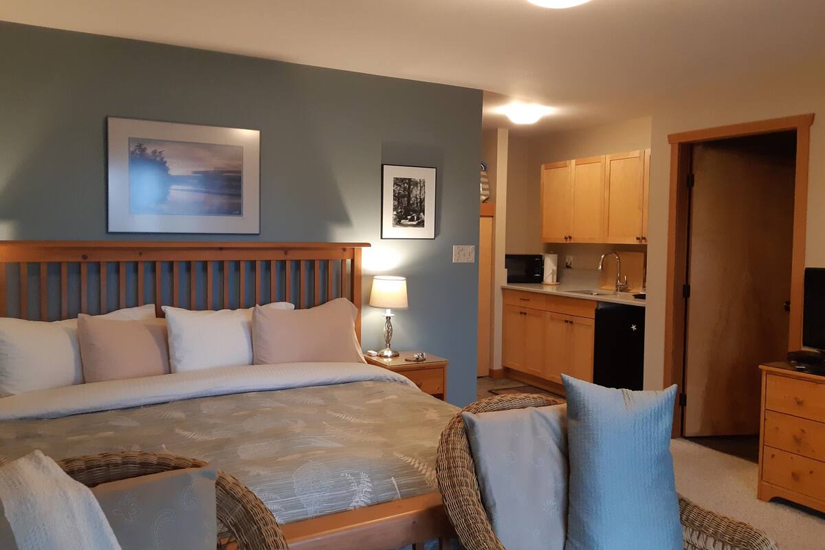 Un dormitorio con una cama grande, una mesita de noche con lámpara, una pequeña mesa con silla y una kitchenette con armarios de madera y microondas.