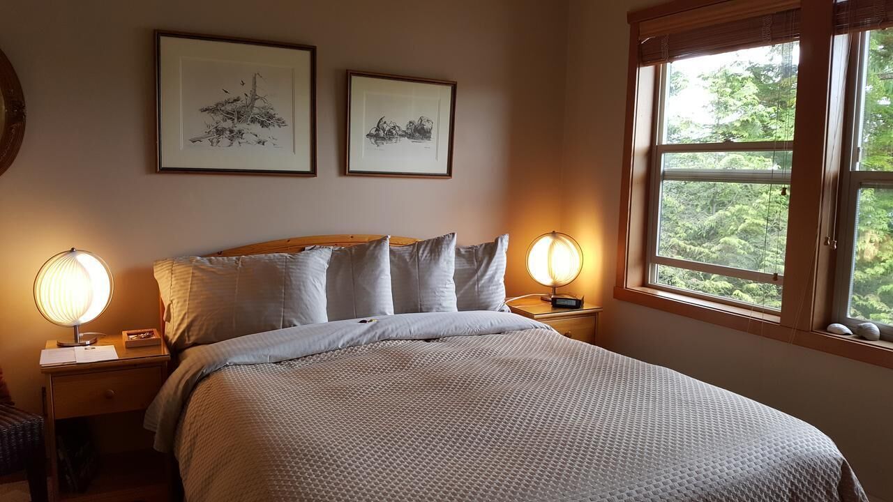 A bedroom with a bed, two bedside tables, a window with a view of trees, and two framed pictures on the wall.