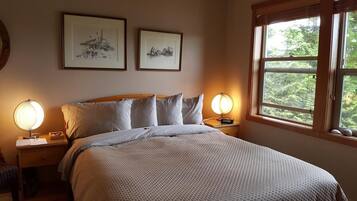 A bedroom with a bed, two bedside tables, a window with a view of trees, and two framed pictures on the wall.