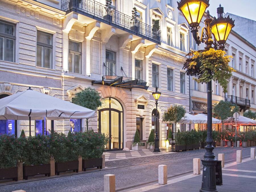 Un bâtiment historique doté d’une entrée imposante, d’un espace extérieur aménagé pour s’asseoir et de réverbères décoratifs.