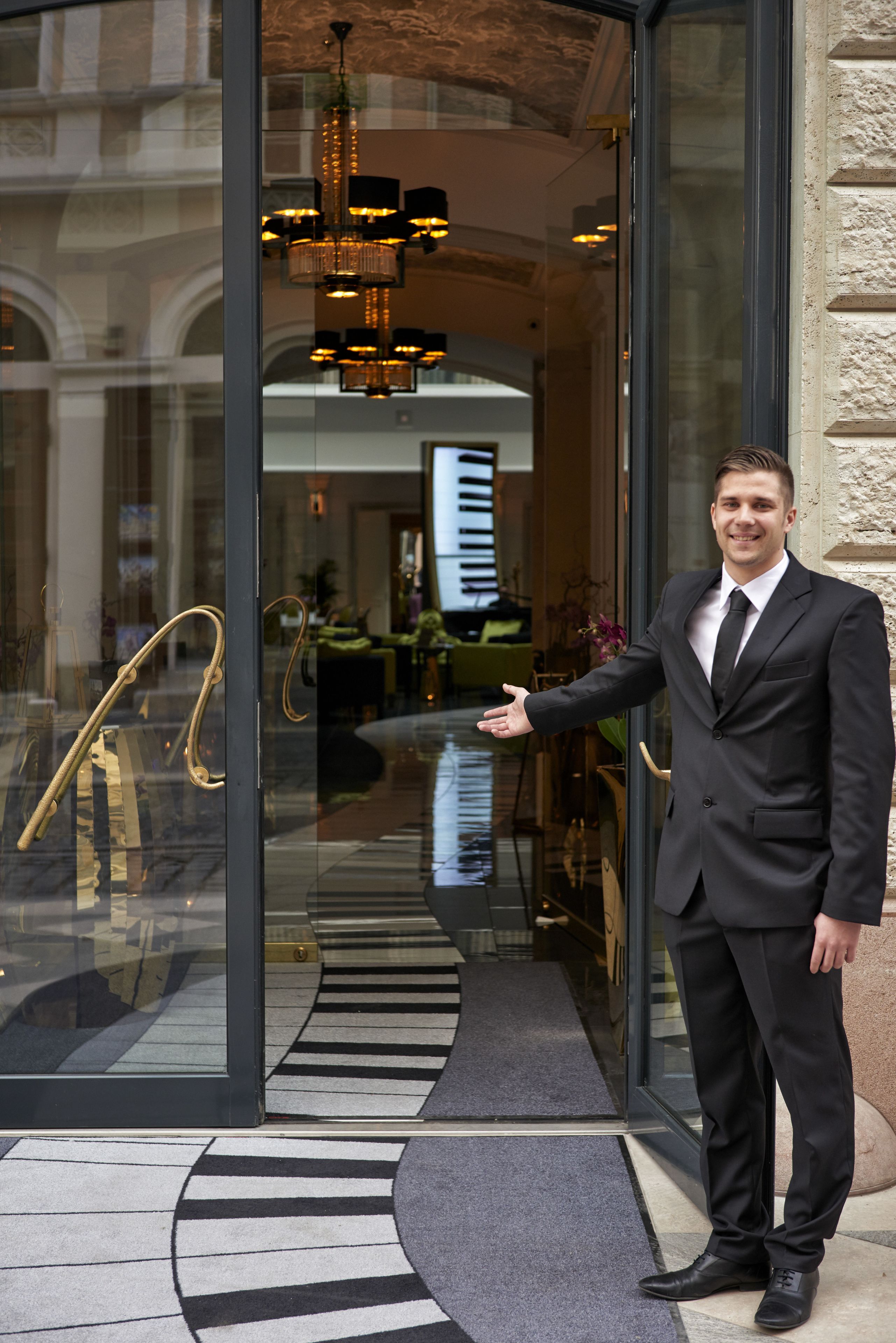 Un hombre con traje de pie en la entrada de un hotel, con puerta de cristal, se distingue el interior con mesas y sillas, y una lámpara de araña.
