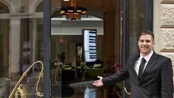 Un hombre con traje de pie en la entrada de un hotel, con puerta de cristal, se distingue el interior con mesas y sillas, y una lámpara de araña.