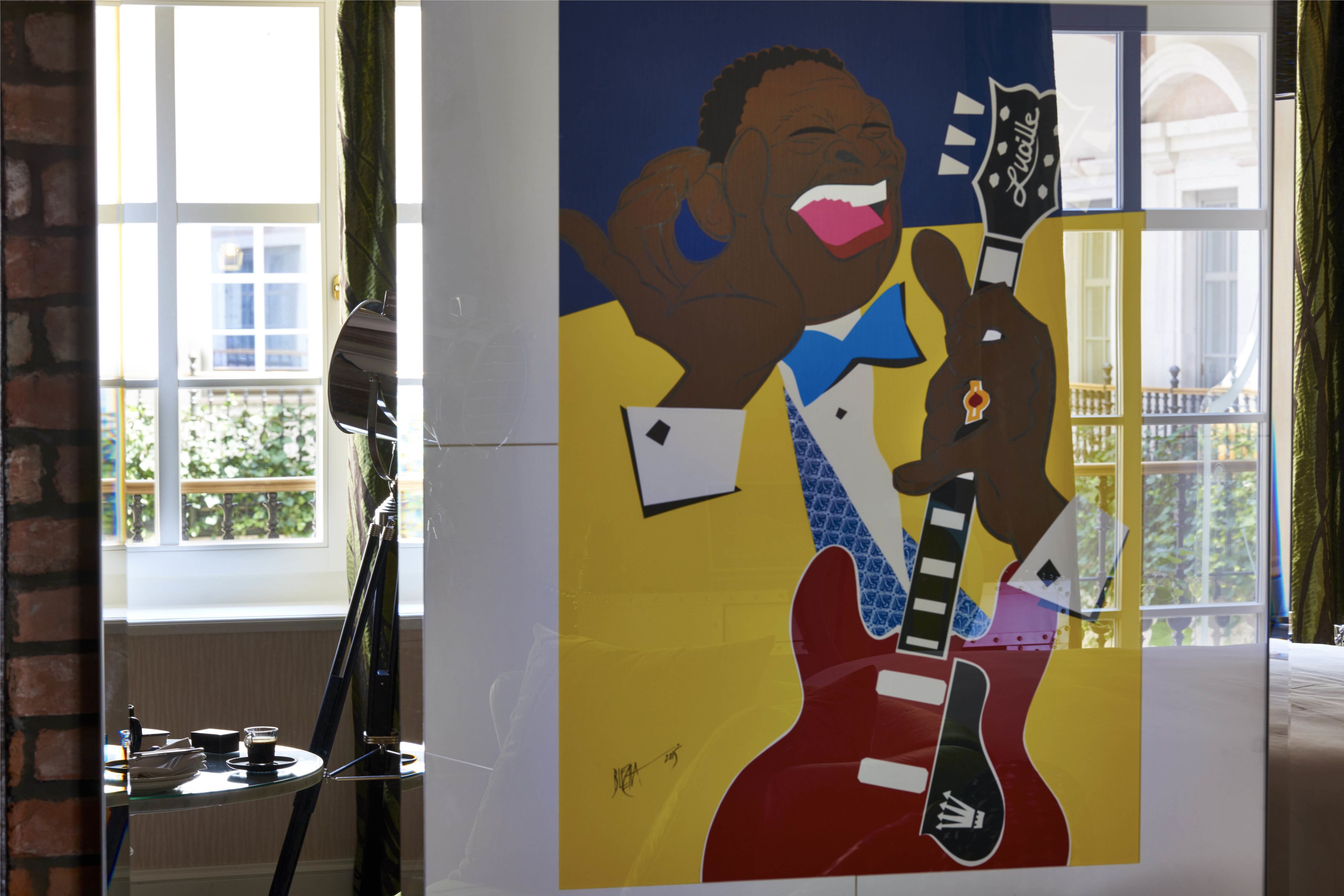 A room with a large window, a painting of a man with a guitar, and a table with a lamp.