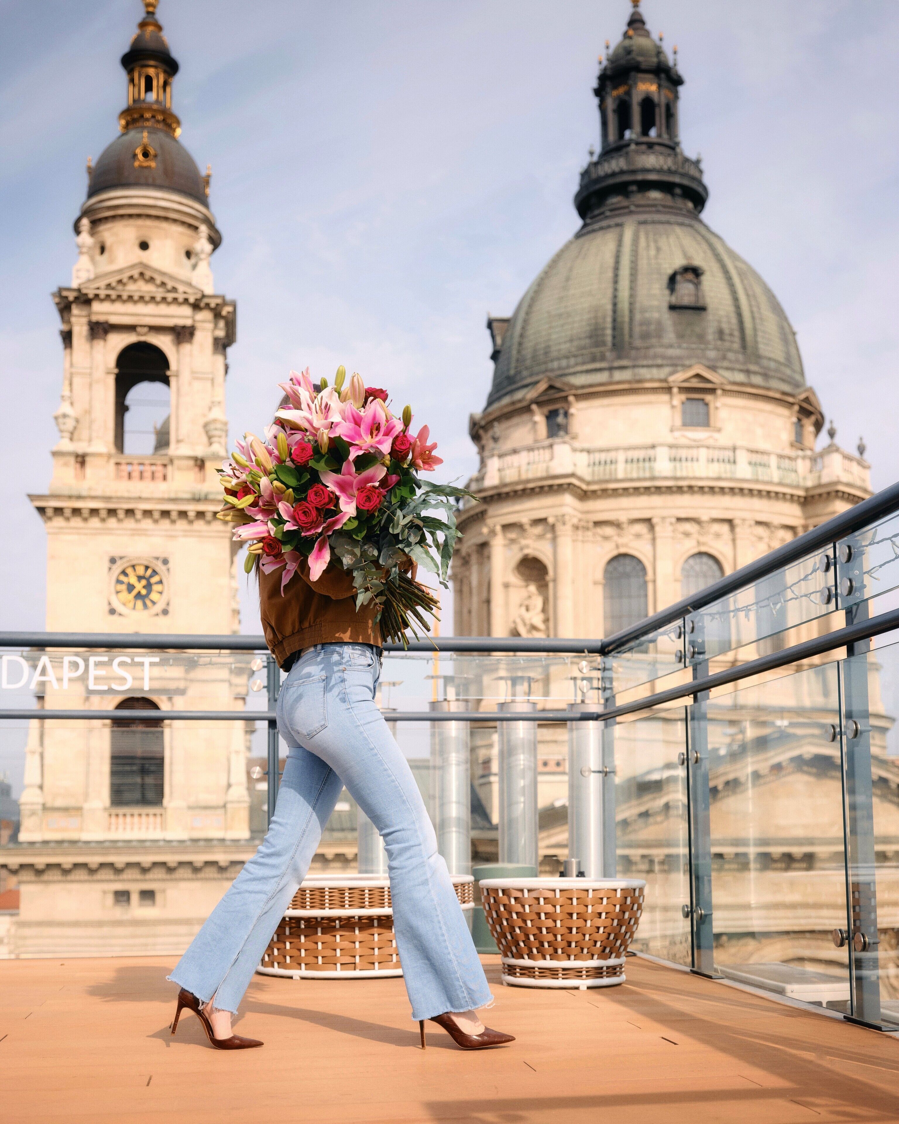 Eine Person geht auf einem Balkon mit einer großen Blumenanordnung, mit Blick auf ein historisches Gebäude mit Kuppeln und einem Uhrturm.