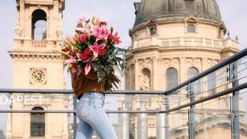 Eine Person geht auf einem Balkon mit einer großen Blumenanordnung, mit Blick auf ein historisches Gebäude mit Kuppeln und einem Uhrturm.