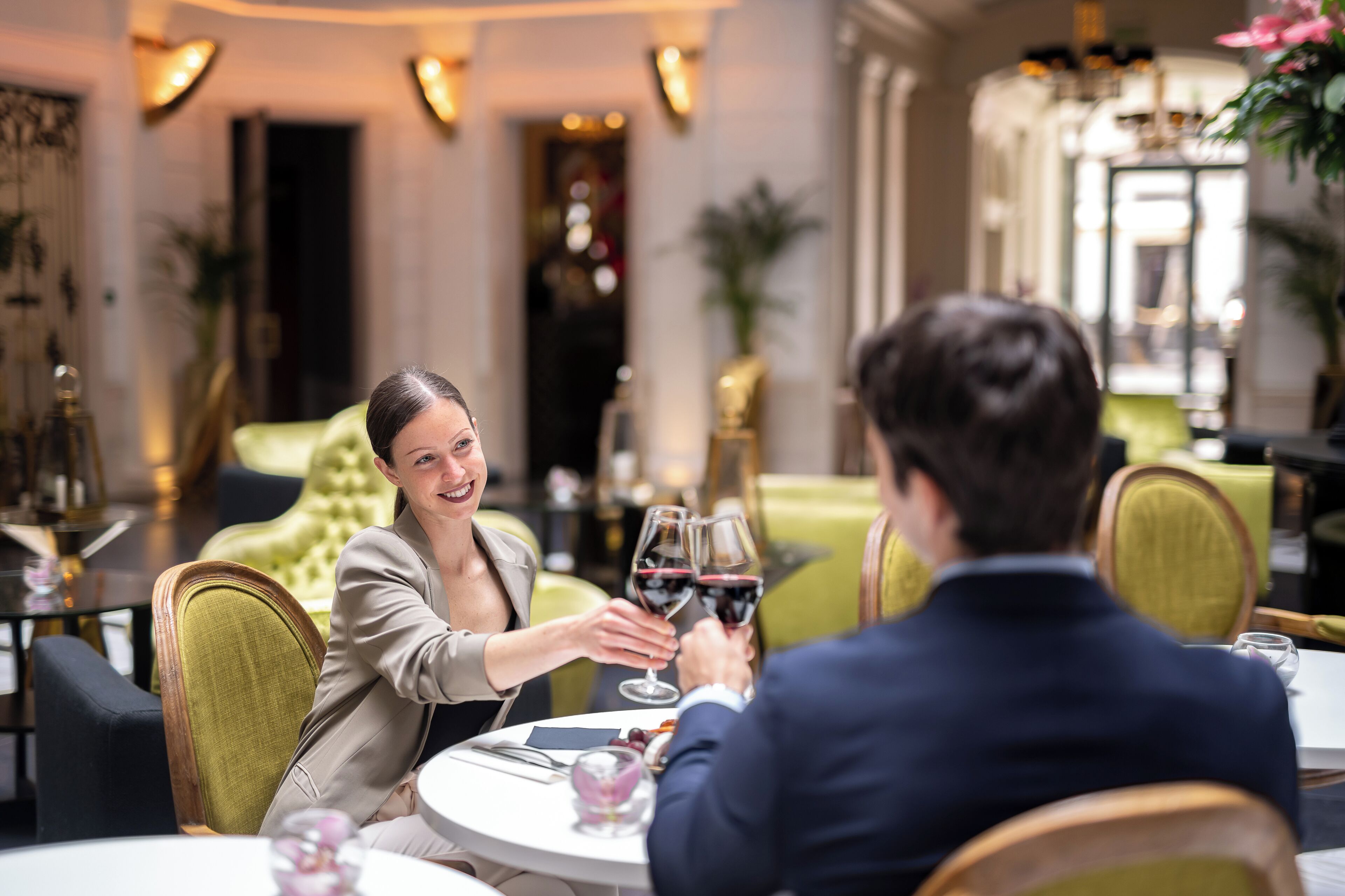 A couple toasting at a round table in a restaurant with a classic interior, including a green upholstered sofa and a chandelier.