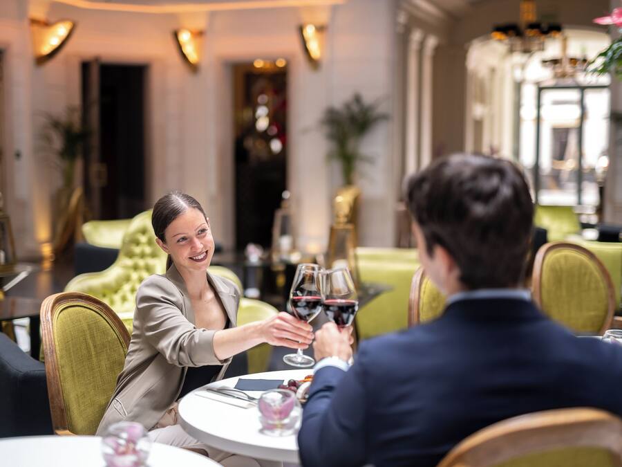 A couple toasting at a round table in a restaurant with a classic interior, including a green upholstered sofa and a chandelier.