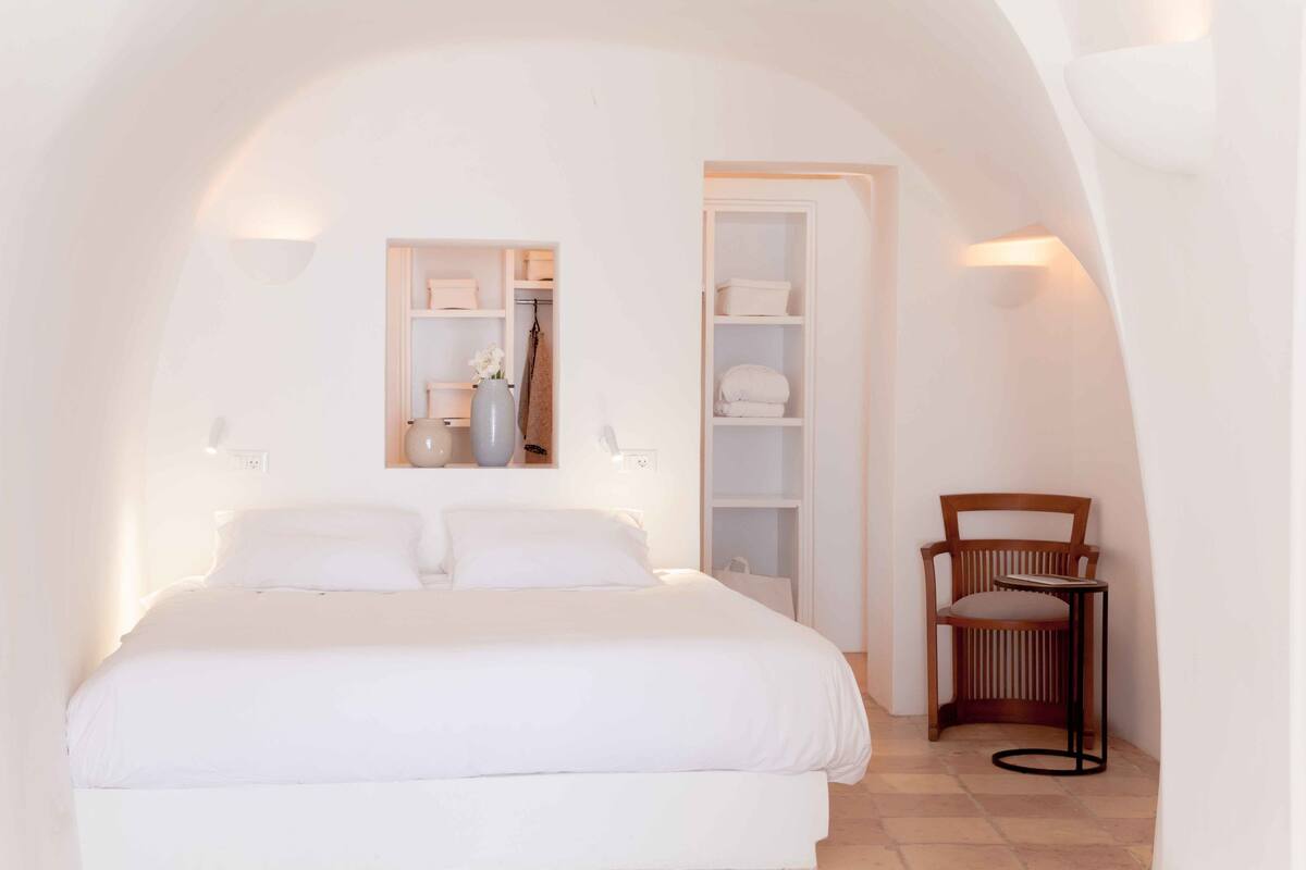 A bedroom with a bed, a wooden chair, a shelf with items, and a white wardrobe.