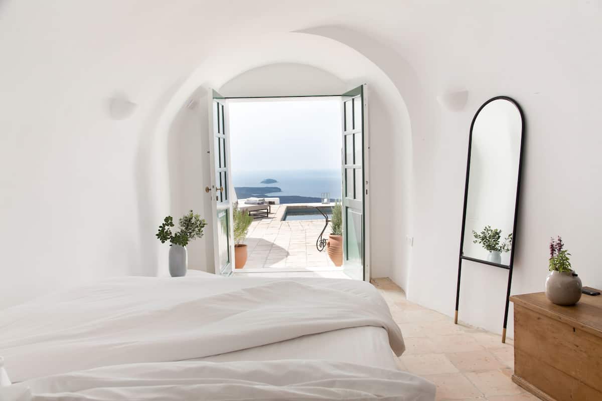 A bedroom with a large bed, an open doorway leading to a patio with a pool and plants, and a mirror on the wall.