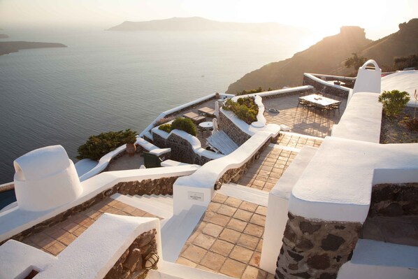 A sunlit terrace with stone steps and white walls overlooking a body of water. - The Vasilicos (Santorini)
