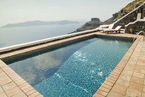 A rectangular pool with clear blue water, surrounded by stone steps and white lounge chairs, overlooking a body of water and distant land. - The Vasilicos (Santorini)