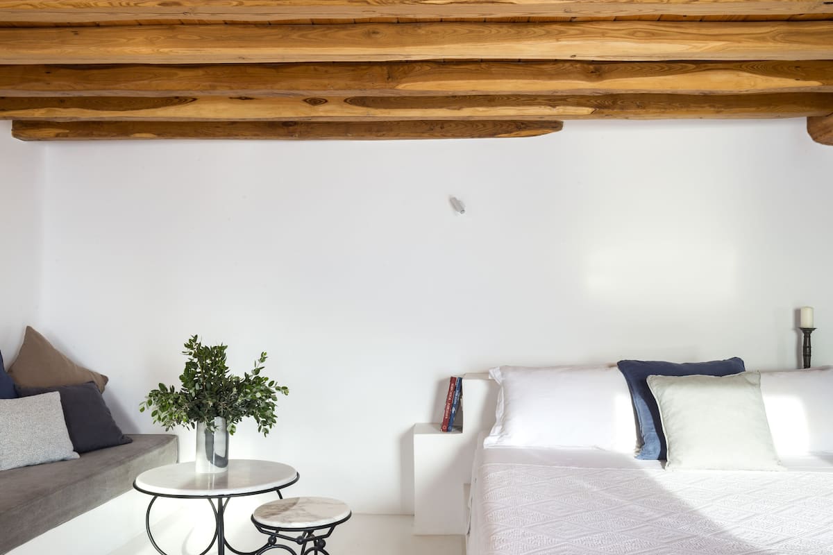 A modern bedroom with a wooden ceiling, a white bed, a grey sofa, a small round table with a plant, and a bedside table with books and a candle.