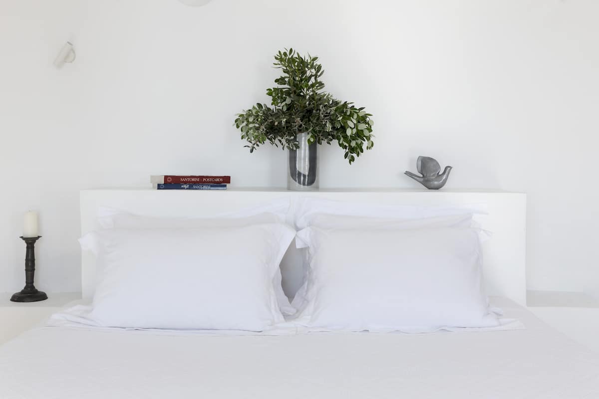 A neatly made bed with white linens and pillows, a small vase with green plants on a white nightstand, a book titled 'SANTORINI POSTCARDS', and a decorative object on the nightstand.