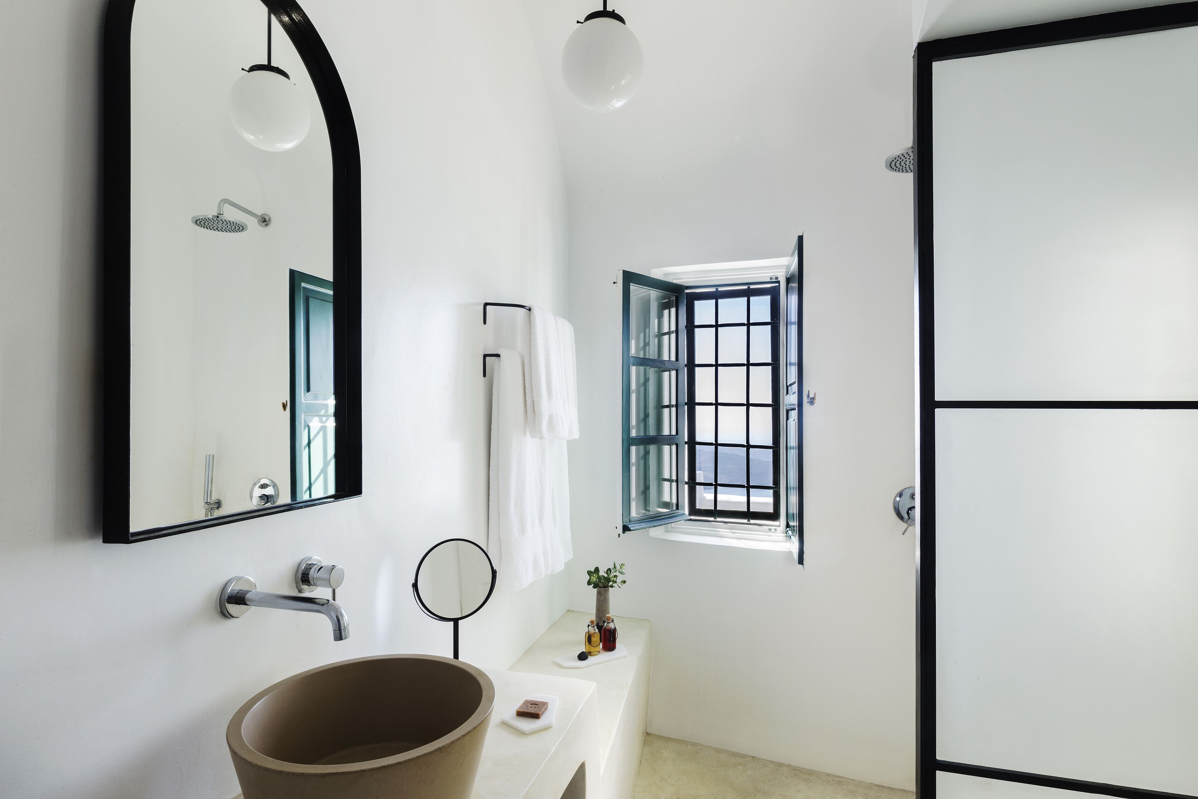 Un baño con un espejo grande, un lavabo redondo, una ducha y una ventana con contraventanas azules.