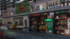 Zona de bar con un letrero que dice 'Bar', estantes con varias botellas y aperitivos, y un enfriador de cerveza Tonia.