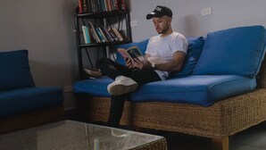 Una persona sentada en un sofá azul leyendo un libro en una habitación con una estantería y una mesa de centro de mimbre.
