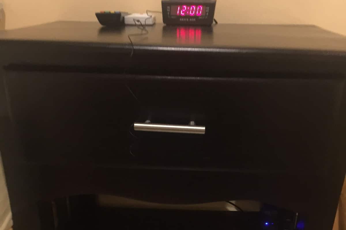 Una mesita de noche negra con un reloj digital que marca las 12:00, una regleta de enchufes y un cajón.