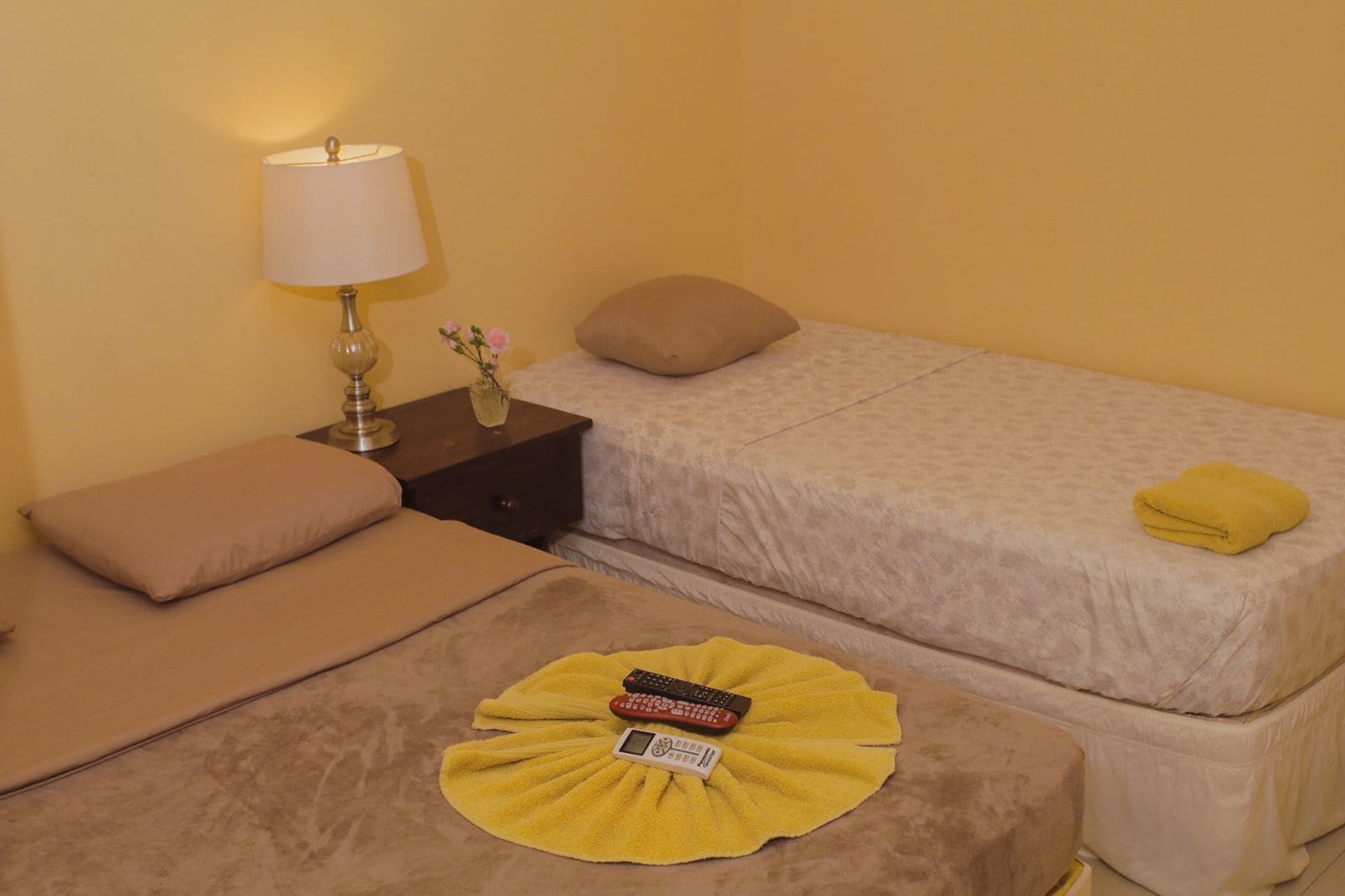 A twin room with two beds, a nightstand with a lamp, a remote control, and a yellow towel.