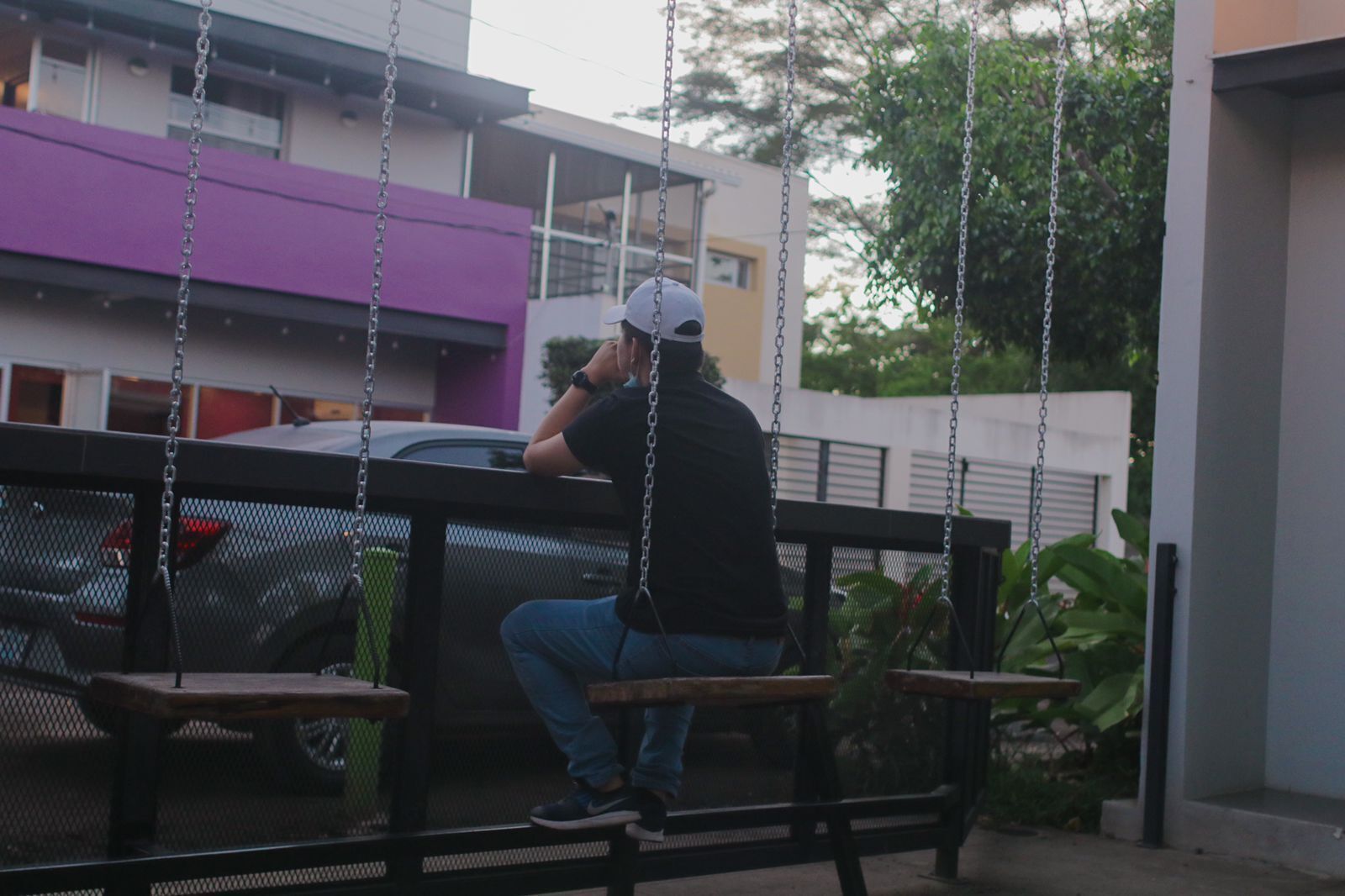 A person sitting on a swing in a fenced outdoor area with a car and buildings in the background.