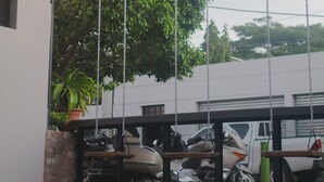 Un área de estar al aire libre con techo, que incluye un conjunto de columpios y guirnaldas de luces, rodeada de coches estacionados y un edificio al fondo.