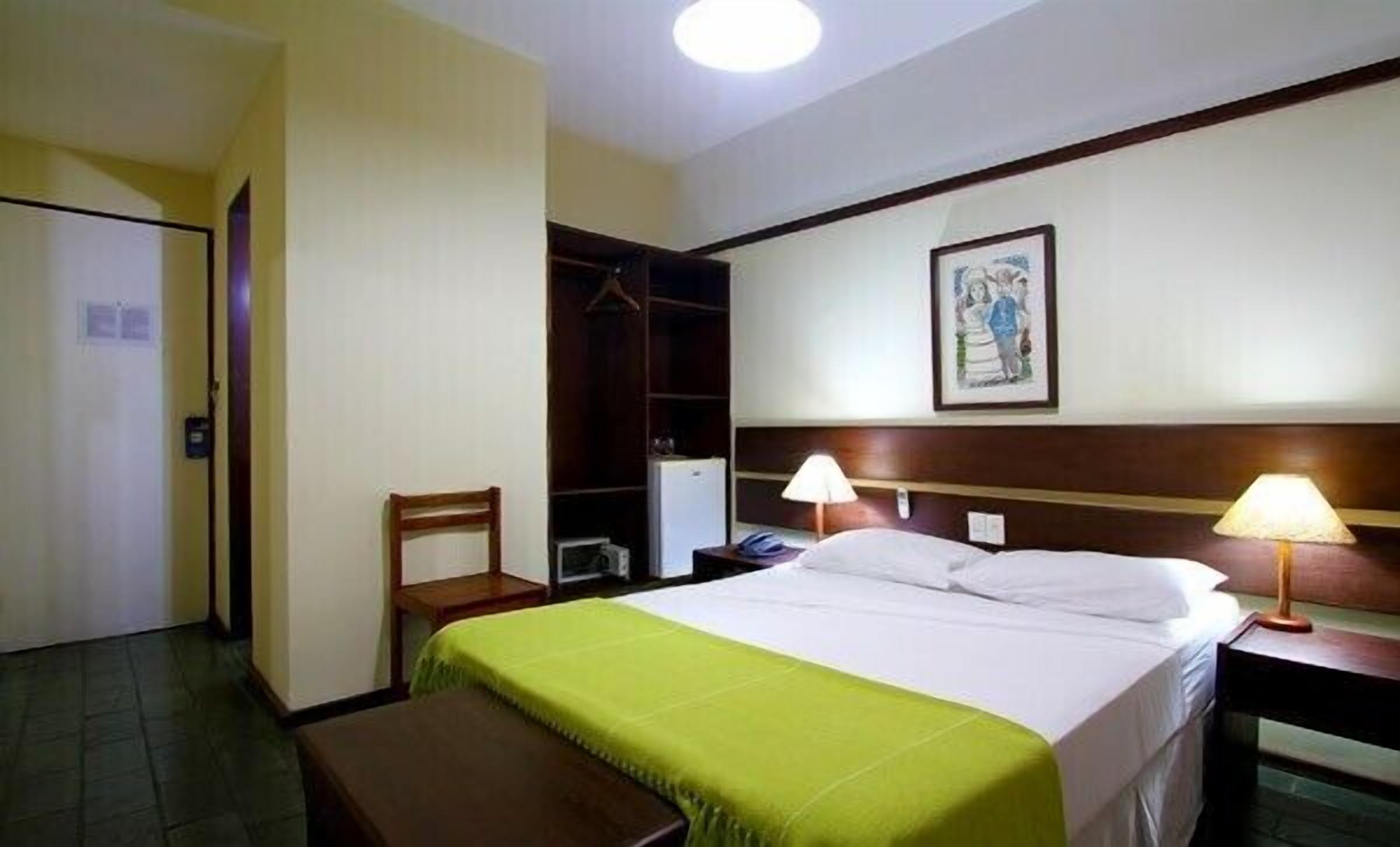 A hotel room with a bed, bedside lamps, a bench, a wardrobe, and a painting on the wall.