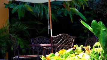 Un patio con una mesa y sillas de metal bajo un paraguas blanco, rodeado de exuberante follaje verde.