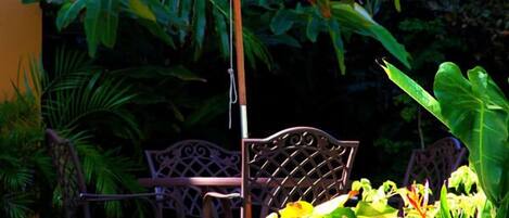 Un patio con una mesa y sillas de metal bajo un paraguas blanco, rodeado de exuberante follaje verde.