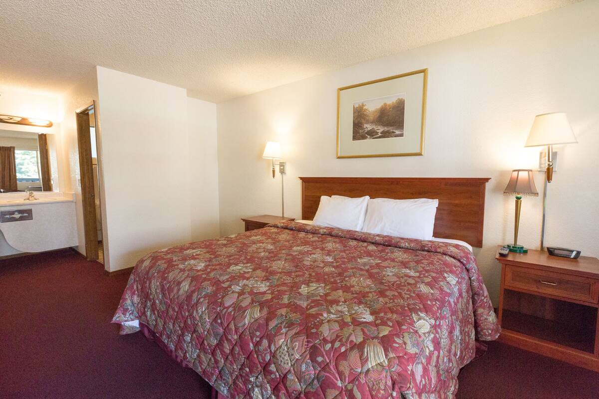 A hotel room with a large bed, a wooden headboard, two bedside lamps, a nightstand, a mirror, and a framed picture on the wall.