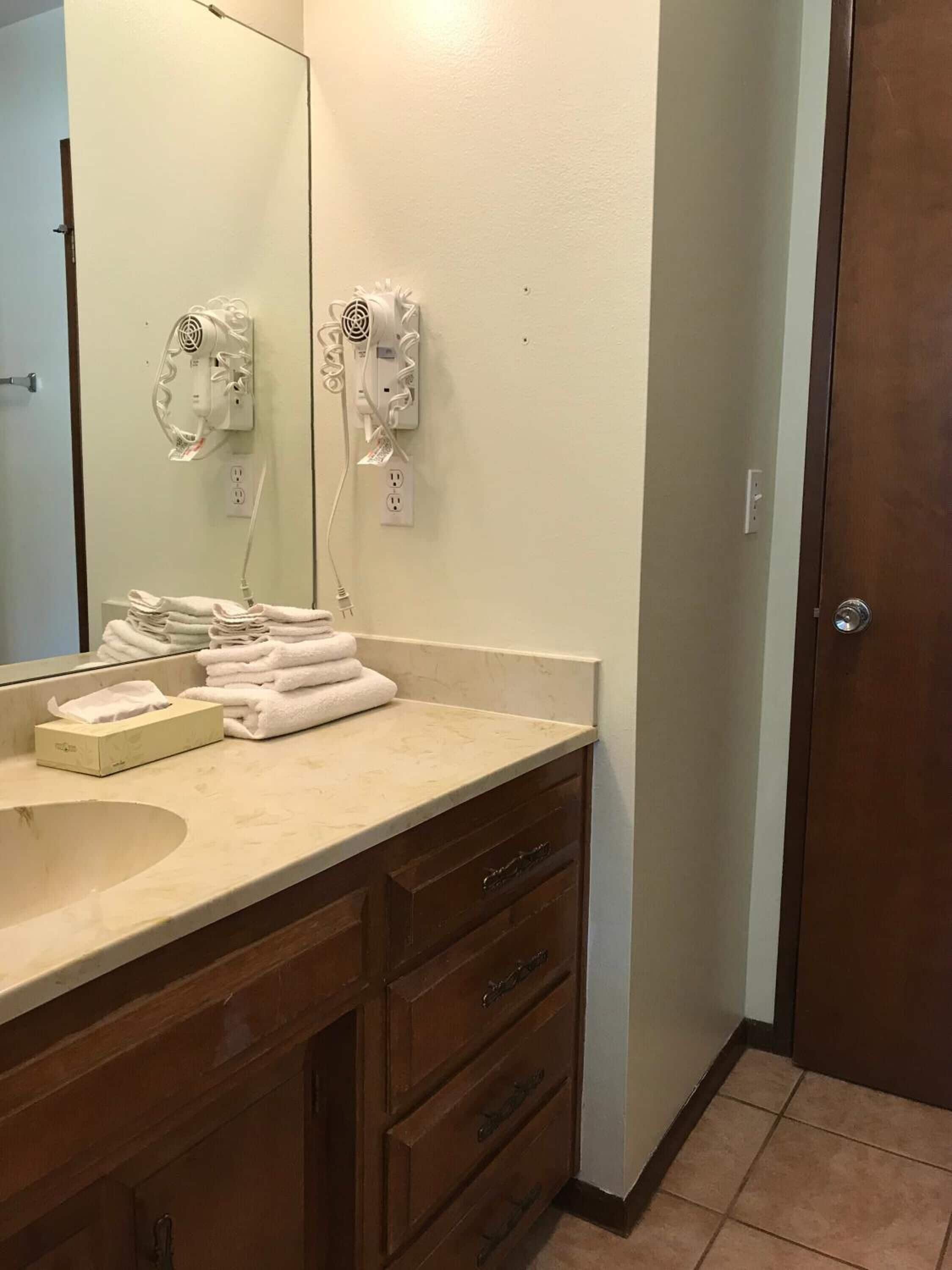 Un baño con lavamanos, espejo, una pila de toallas y un secador de pelo montado en la pared.