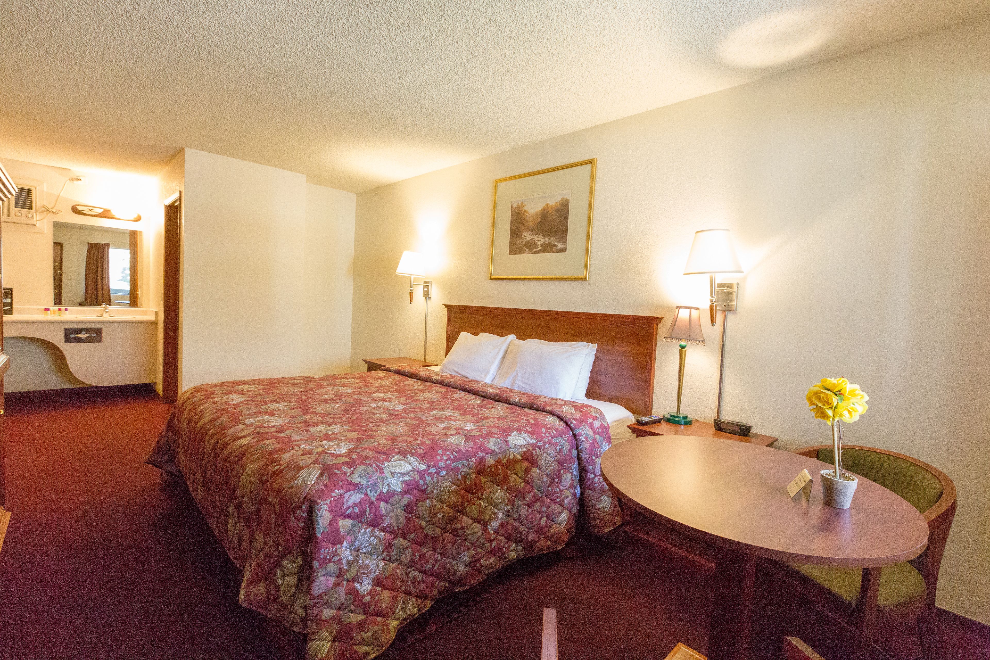 A hotel room with a bed, a desk, a chair, and a small table with a flower vase.