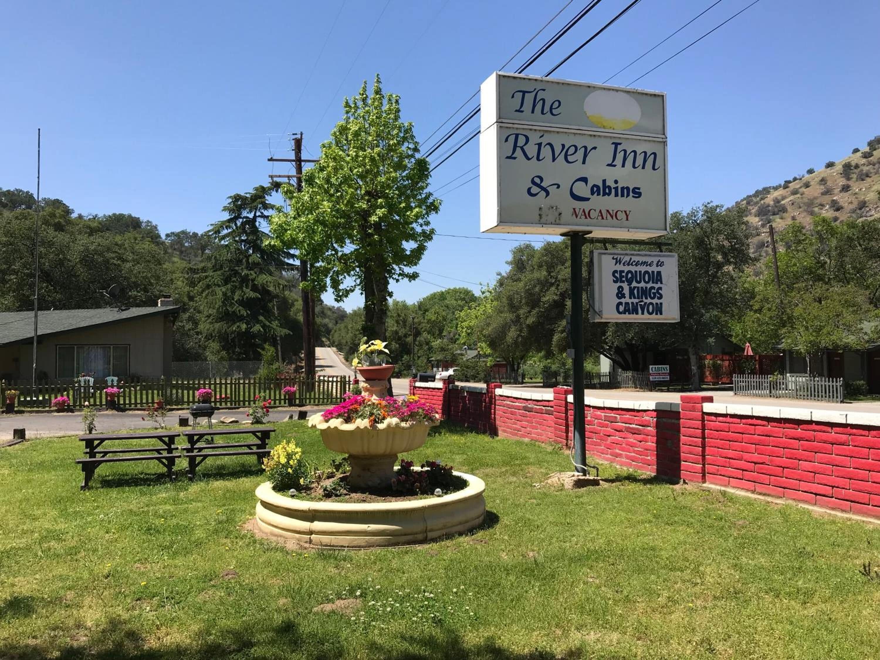 The River Inn & Cabins sign with a 'VACANCY' notice, surrounded by a red brick wall, a picnic table, and a flower bed.