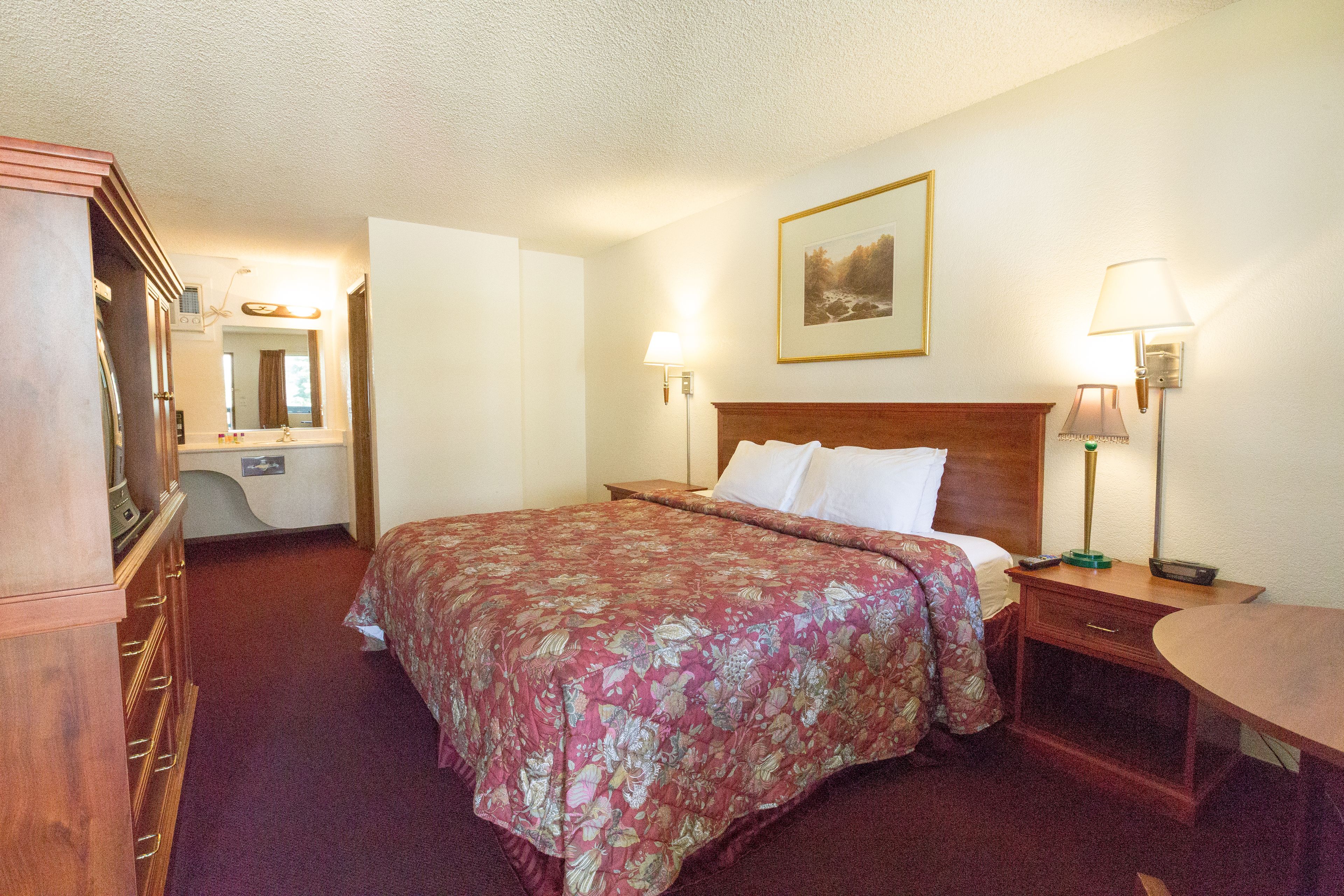 A hotel room with a large bed, a wooden dresser, a desk, a chair, a lamp, a bedside table, and a framed picture on the wall.