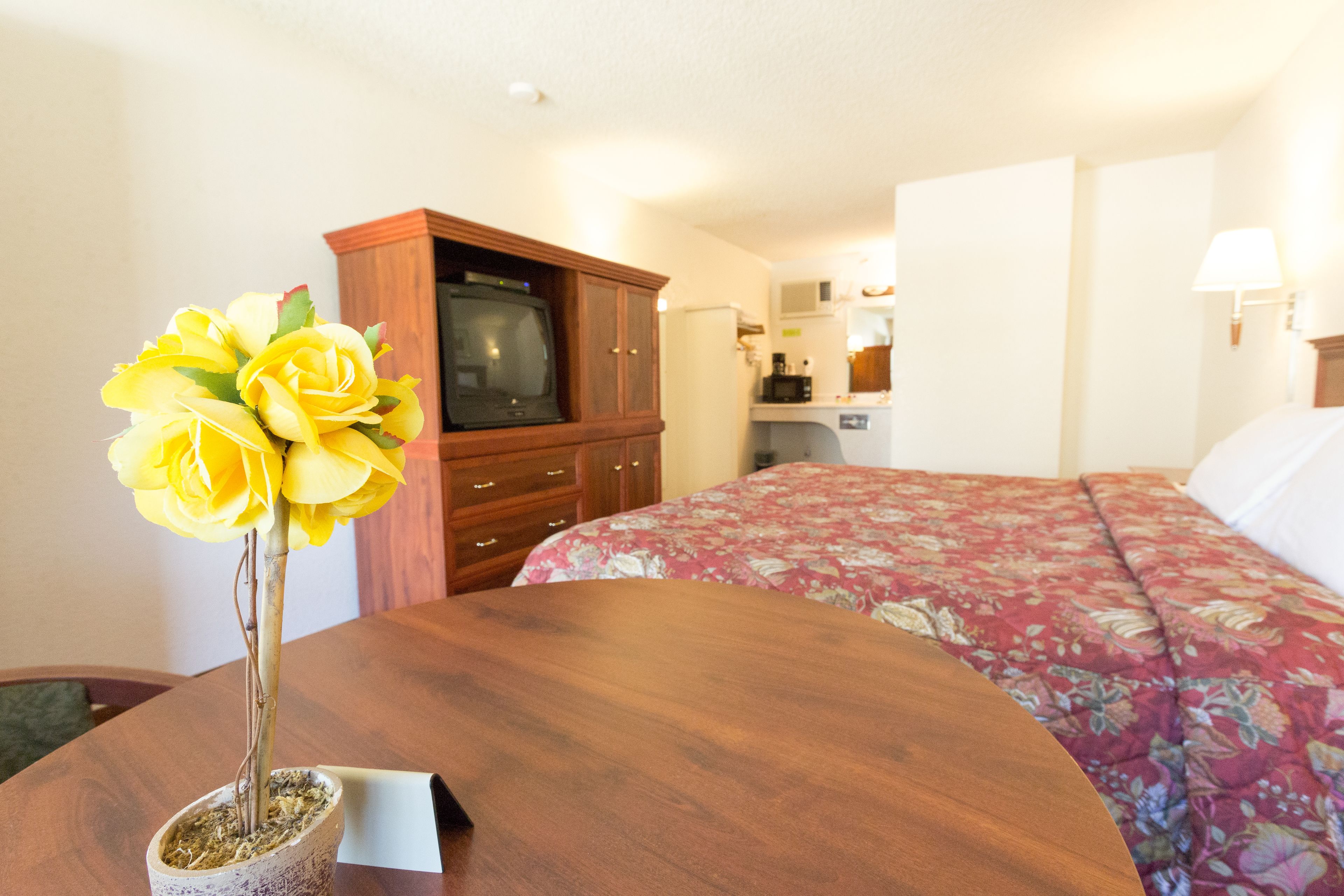 A hotel room with a wooden dining table, a floral bedspread, a television, and a kitchenette in the background.
