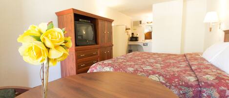 A hotel room with a wooden dining table, a floral bedspread, a television, and a kitchenette in the background.