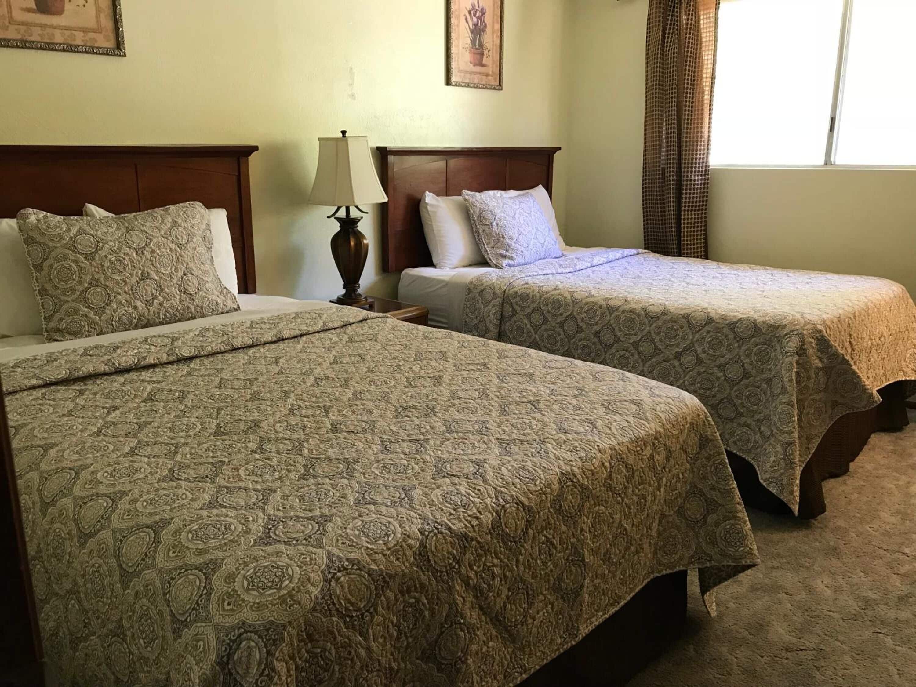 A hotel room with two beds, each with a patterned bedspread, a nightstand with a lamp, and a window with curtains.