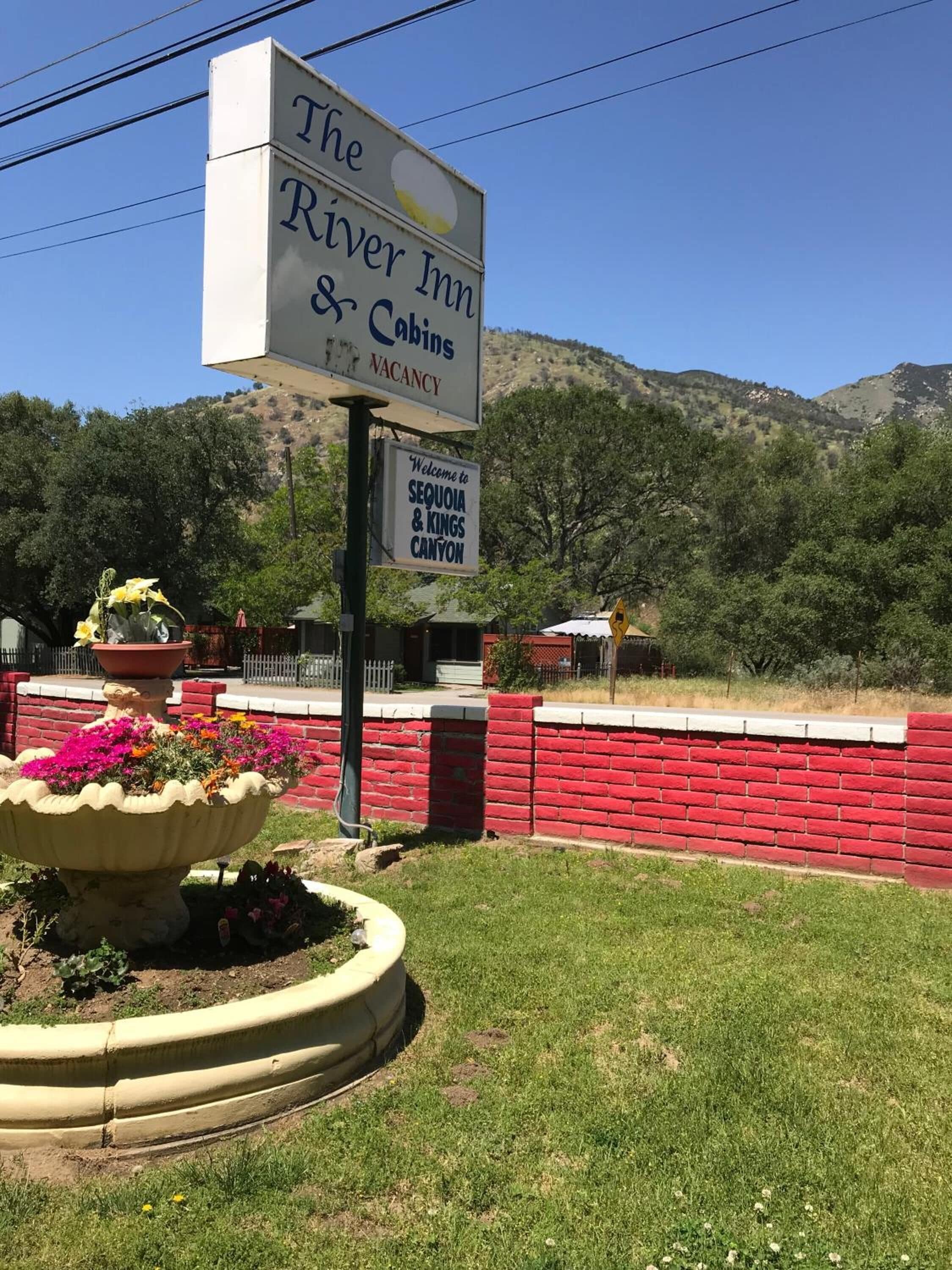 A sign for The River Inn & Cabins with a vacancy notice, a welcome sign to Sequoia & Kings Canyon, and a flower bed.