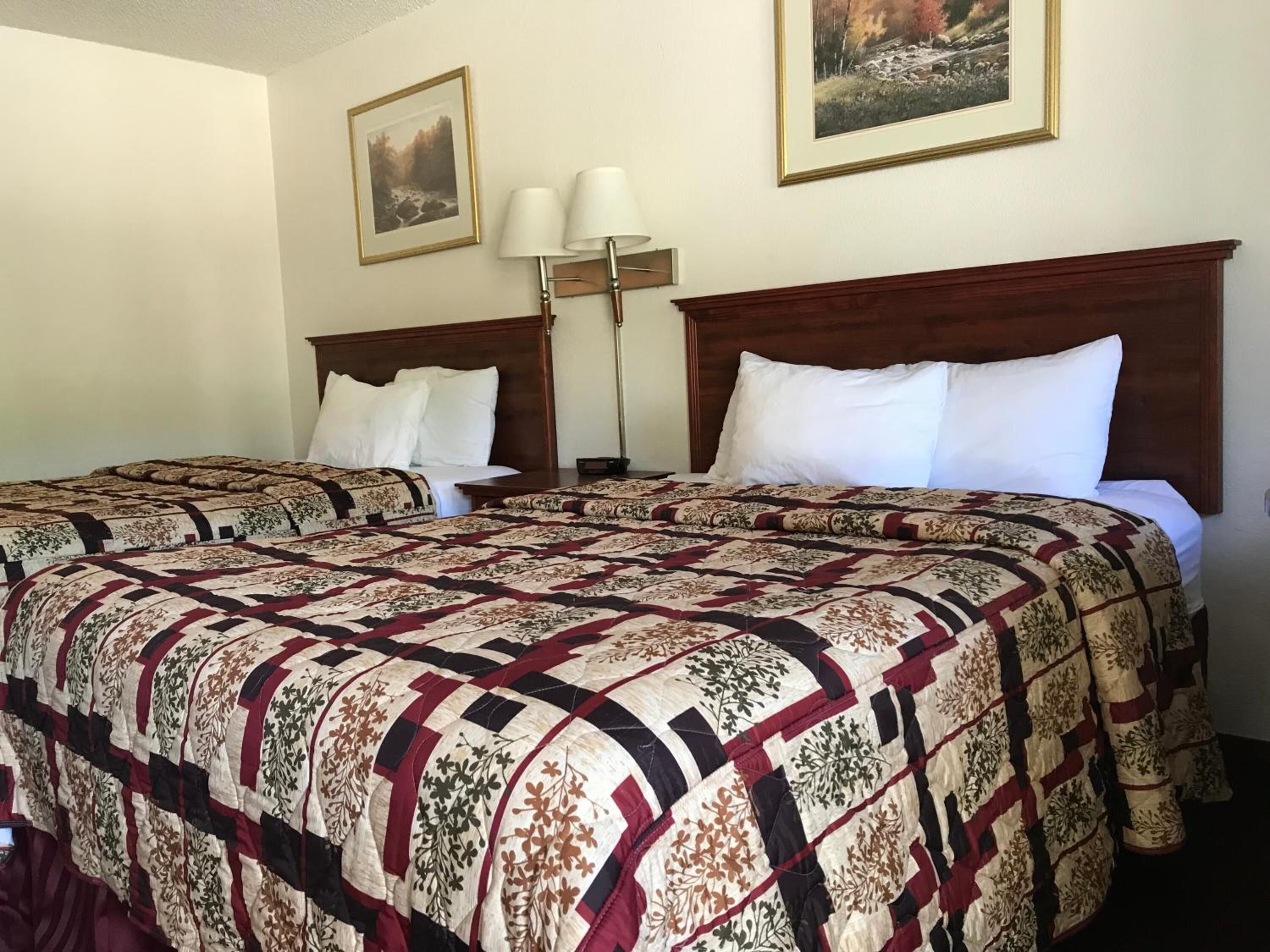 A hotel room with two beds, a wooden headboard, a nightstand, a lamp, and two framed pictures on the wall.