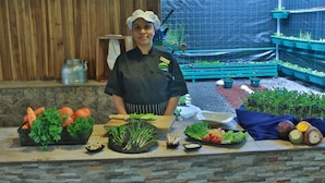 Un chef se tient derrière un comptoir, devant une sélection de légumes frais, d’herbes aromatiques et de divers autres produits alimentaires.