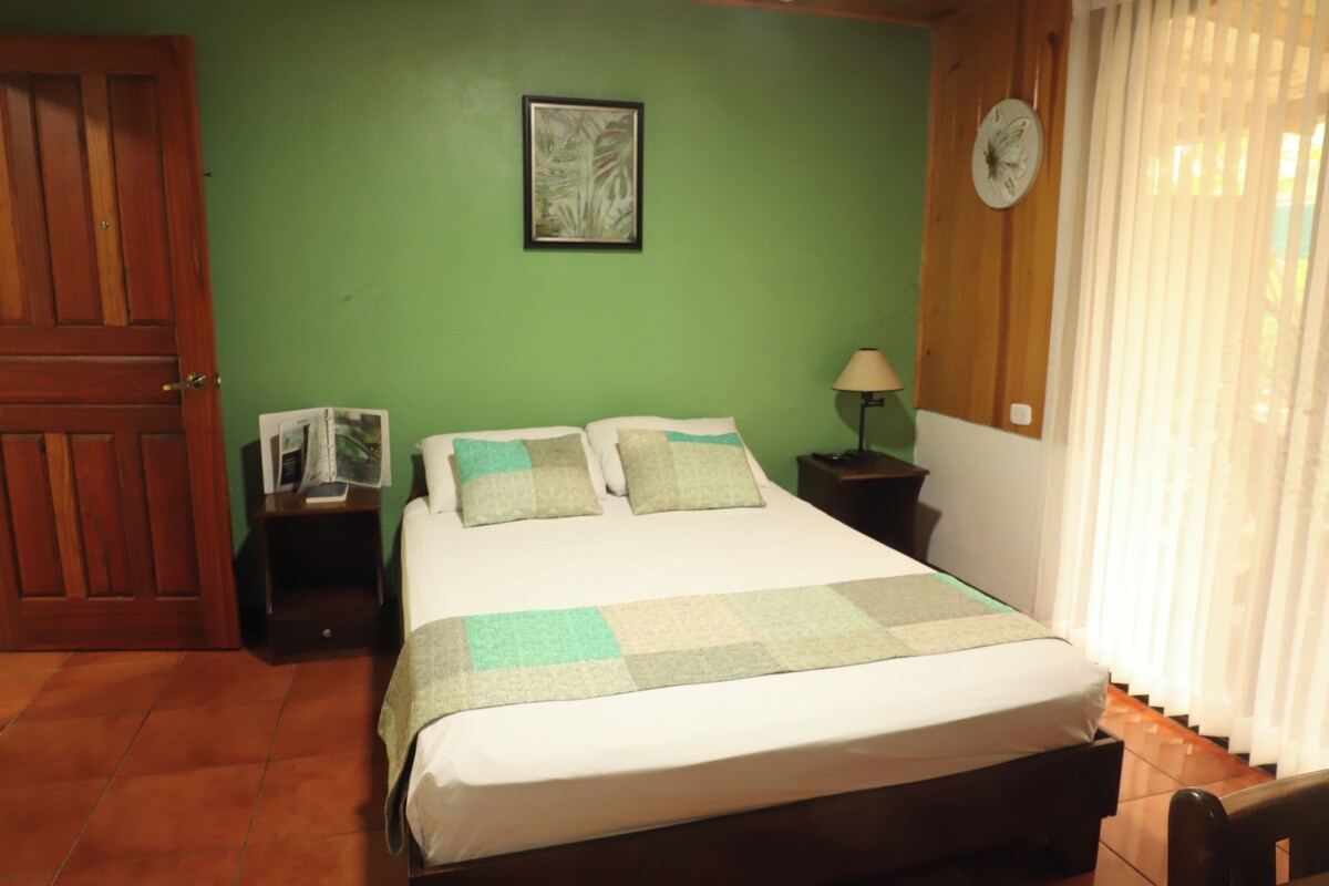 Una habitación con una cama, una mesita de noche, un reloj de pared, un cuadro enmarcado y una puerta.