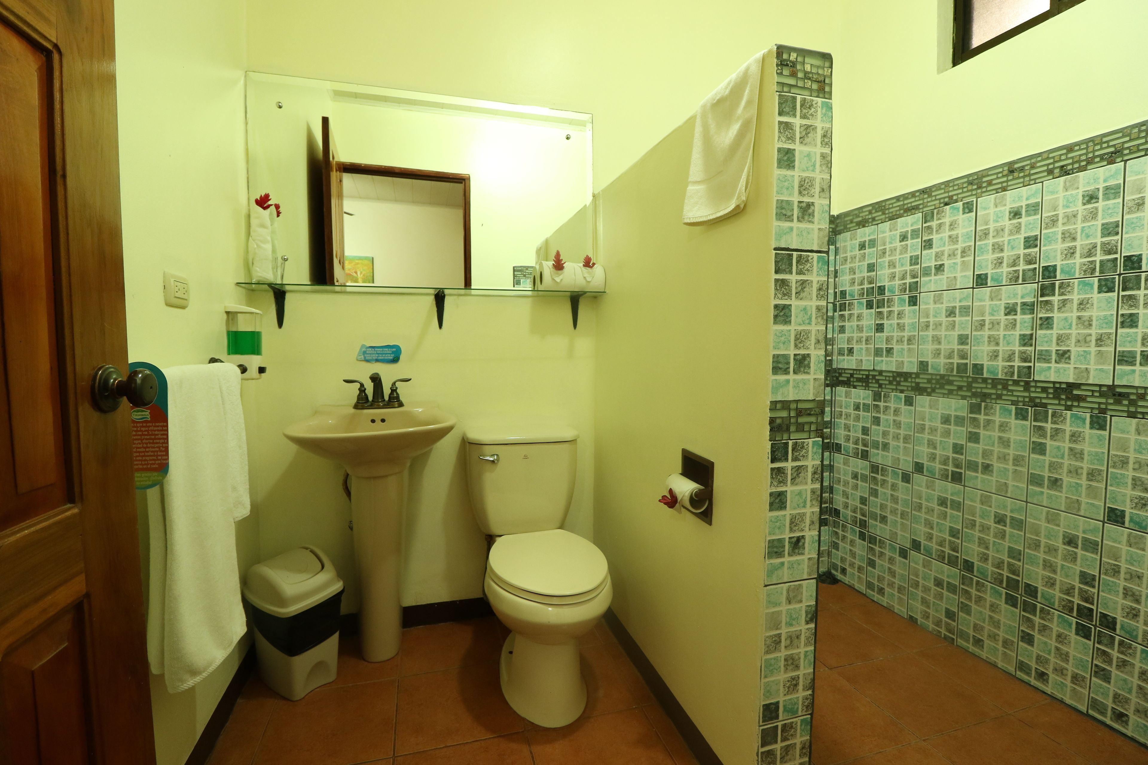 A bathroom with a toilet, sink, and mirror. There is a towel hanging on the wall and a trash bin.