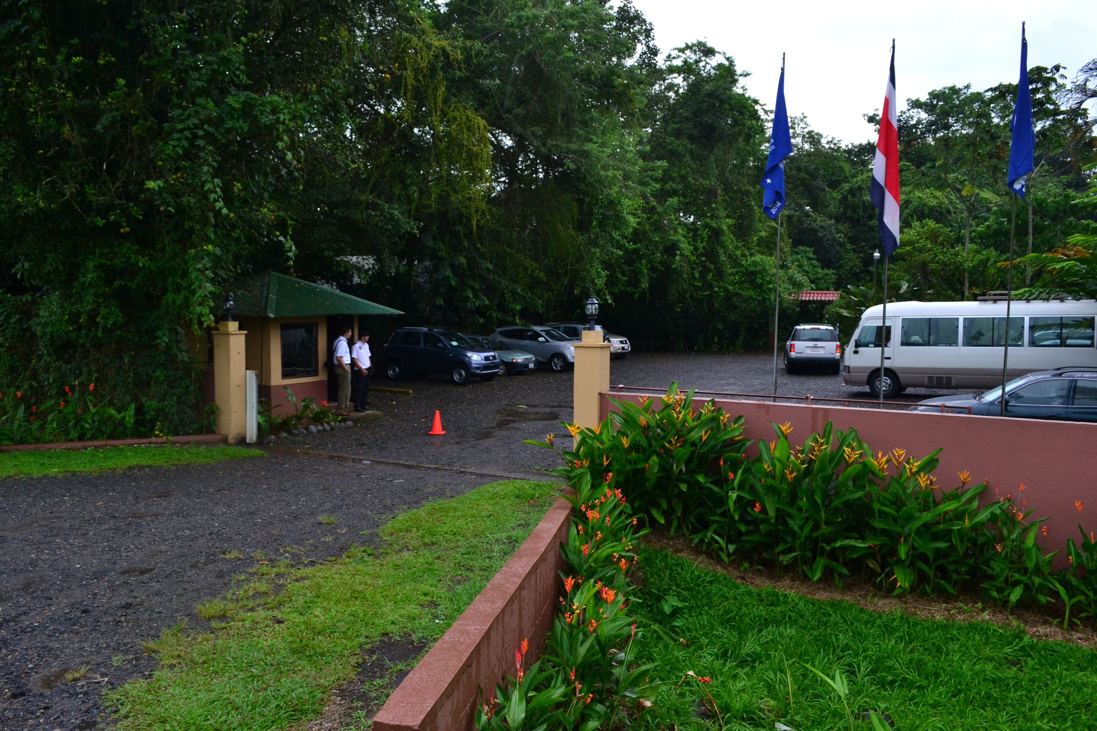 A parking area with cars and a van, surrounded by greenery and flags.