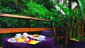Une table dressée avec des aliments pour le petit-déjeuner, notamment des toasts, des œufs et des fruits, sur une nappe violette, à l’extérieur, avec une rambarde en bois et une végétation luxuriante en arrière-plan.