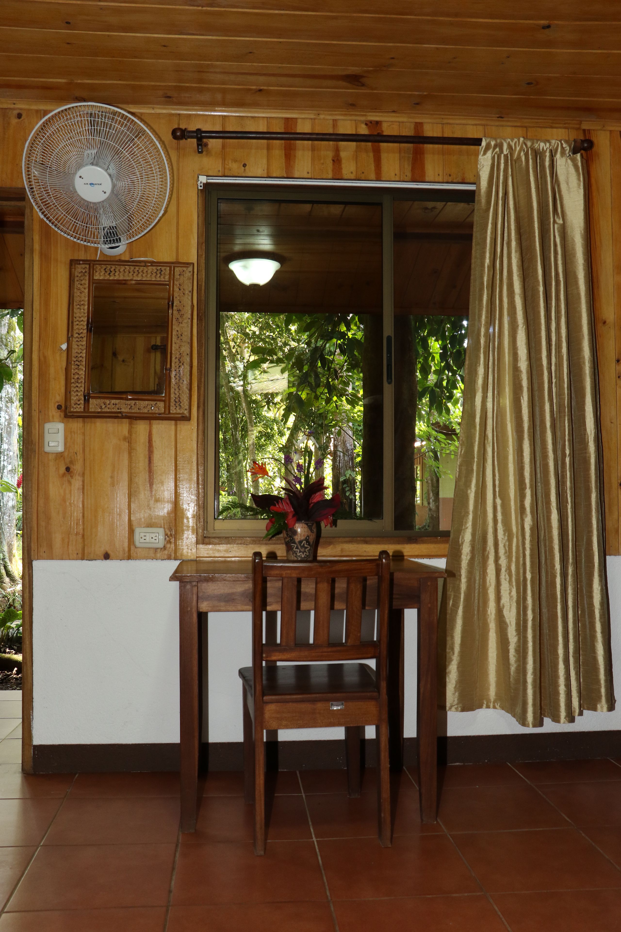 A wooden room with a dining table, chairs, a fan, a window with a curtain, and a mirror on the wall.