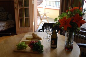 Una mesa con queso, uvas y una botella de vino, un florero con flores, y vista a un balcón con sillas.
