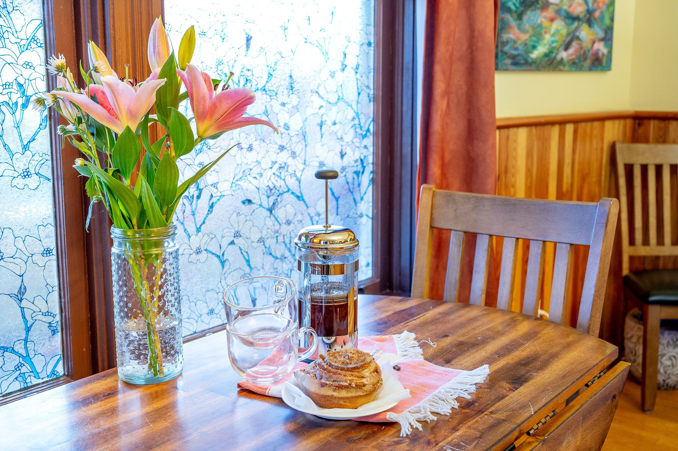 A wooden table with a glass jar of flowers, a glass cup, and a coffee press.