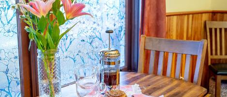 A wooden table with a glass jar of flowers, a glass cup, and a coffee press.