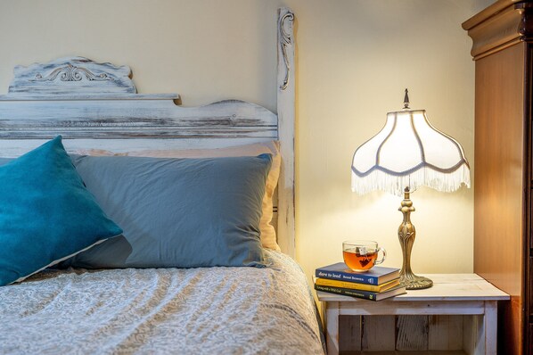 Una cama bien hecha con un almohadón azul, una mesita de noche con una lámpara y una pila de libros.