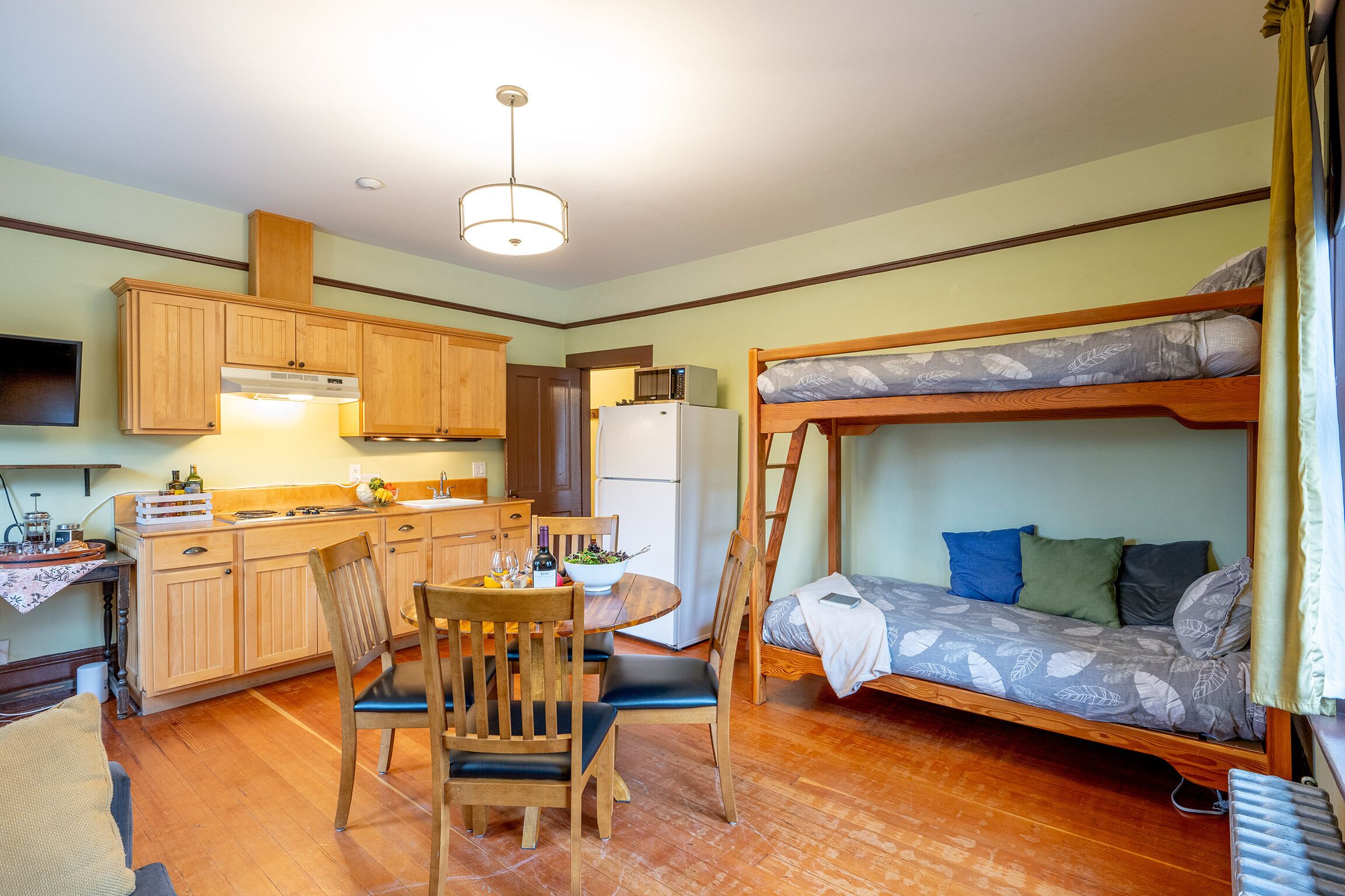 A compact kitchen with wooden cabinets, a dining table with chairs, a bunk bed with patterned bedding, and a ceiling light.